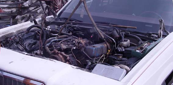 Engine bay of a white sedan with hood open, showing a grimy V8 engine with hoses and wiring
