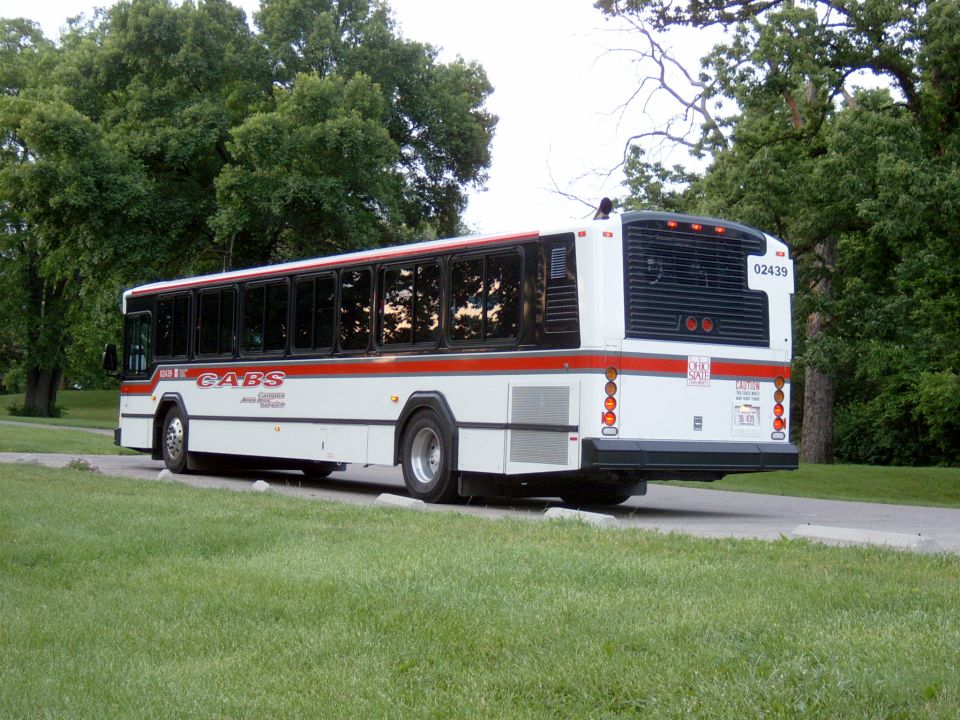 White CABS transit bus with red stripe, rear quarter view parked among green trees