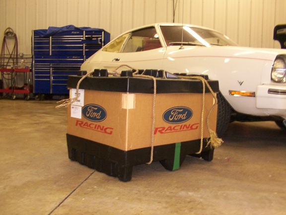 Ford Racing cardboard box roped to black crate in front of a white coupe in a garage.