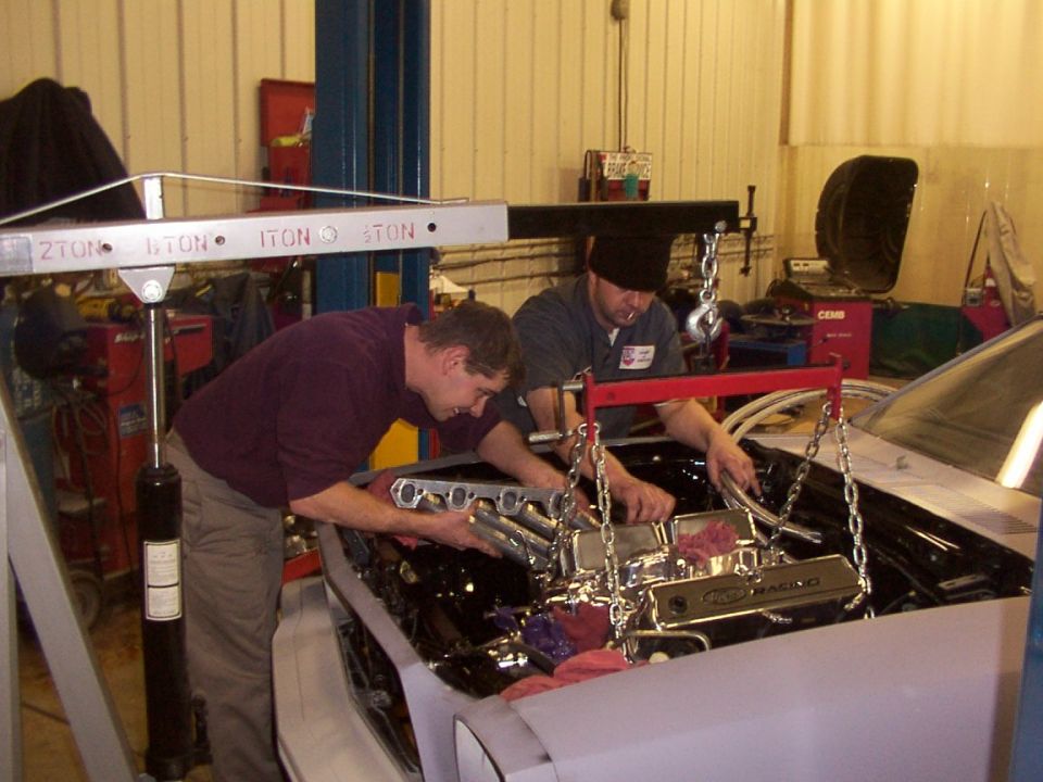 Two men installing a V8 engine with Ford Racing valve covers into a gray car using an engine hoist in a garage