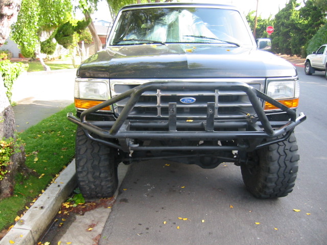 Front of dark Ford Bronco with black tubular bumper on large off-road tires, parked at curb