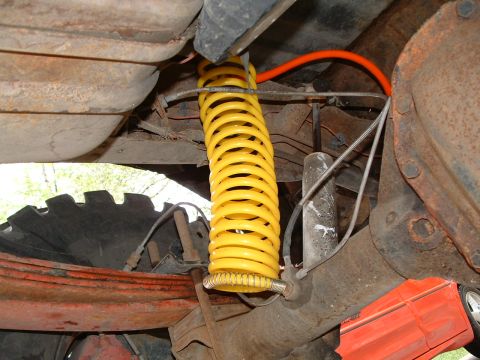 1979 Ford Bronco - Underbody view of a solid-axle front suspension showing a bright yellow coil spring seated on the axle perch, with a smaller silver pigtail…