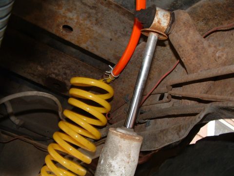 1979 Ford Bronco - Close-up under a truck showing a bright yellow lift coil spring beside a white-bodied shock absorber with an exposed chrome shaft and rubbe…