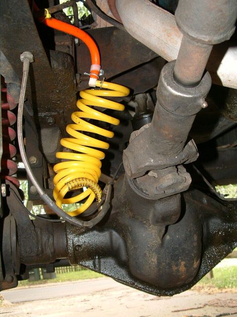 1979 Ford Bronco - Underside close-up of a solid front axle with a yellow coiled brake-hose extension clamped to the frame bracket and an orange braided line…