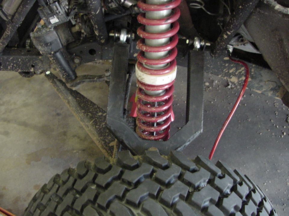 Coilover shock with red spring mounted in a fabricated link bracket above a mud tire, underbody