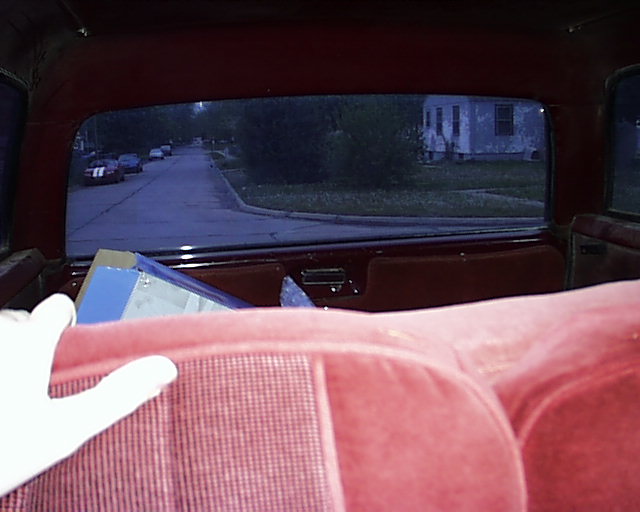View out the rear window of a vehicle over red velour rear seats onto a residential street