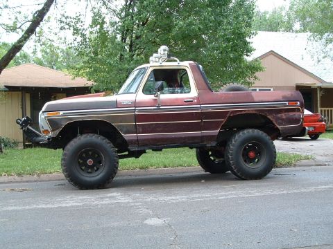 1979 Ford Bronco - Driver-side profile of a lifted maroon-over-tan full-size Ford Bronco with the hardtop removed, parked on a residential street. It rides on…