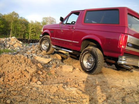 1994 Ford Bronco - A red full-size Ford Bronco flexing over a rock pile on a dirt lot, shot low from the driver's side rear quarter with the front wheel climb…