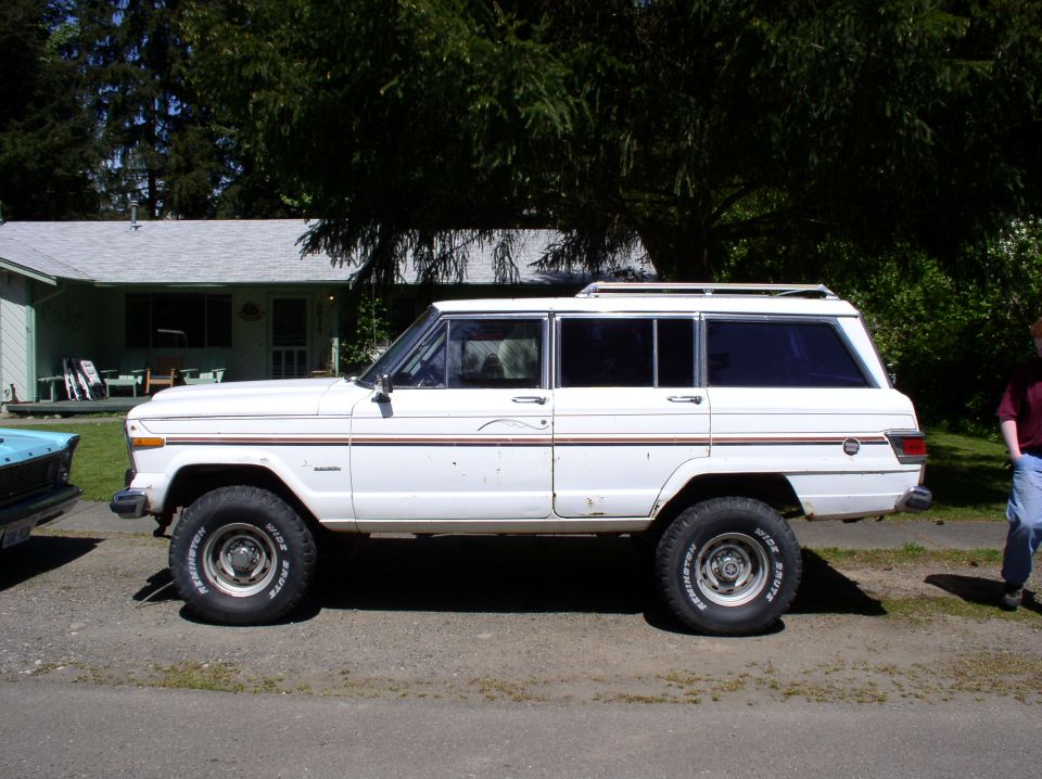 Side view of a white four-door Jeep SUV with roof rack and Remington Wide Brute tires, parked on gravel
