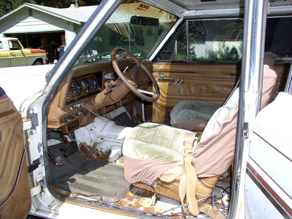 Open driver's door of white Jeep SUV showing worn tan interior, rusty bare floor, and beige seat covers