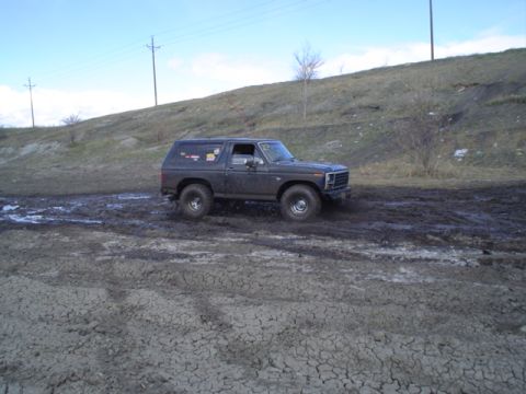 1981 Ford Bronco - A dark gray Ford Bronco navigates a muddy terrain, with its tires partially submerged in the mire.