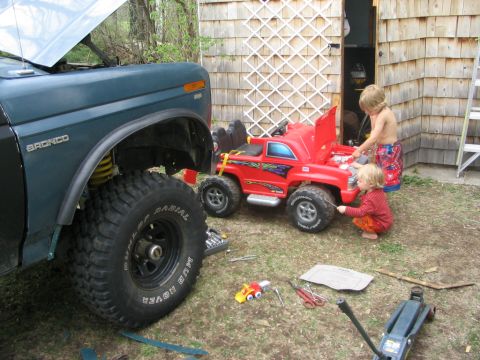 1986 Ford Bronco - Two children work on a red toy truck near a 1986 Ford Bronco with its hood open.