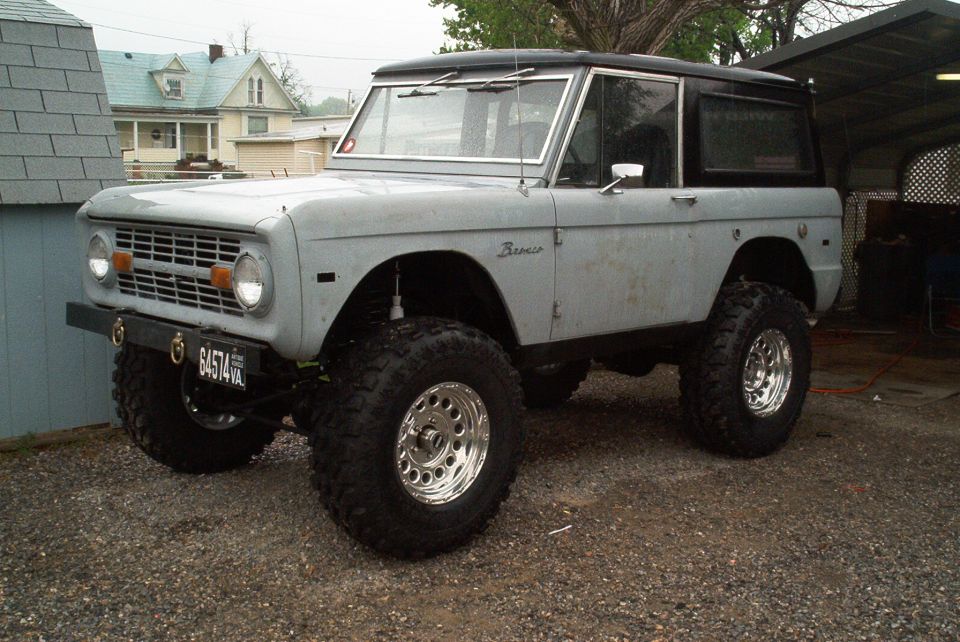 Gray early Ford Bronco with black top, lifted on big tires, parked on gravel by a teal shed.