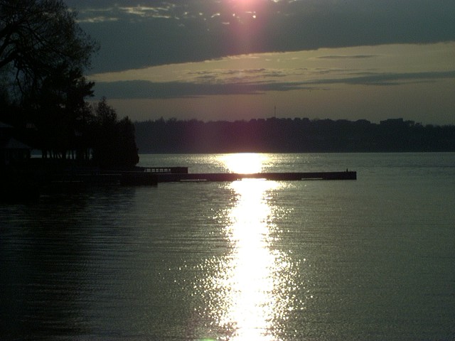 Sunset over a lake with a dock extending into the water. Trees and a distant treeline are visible