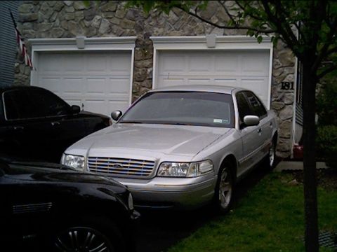2002 Ford Crown Victoria - A silver sedan is parked in front of a house with two garage doors, with another car partially visible.