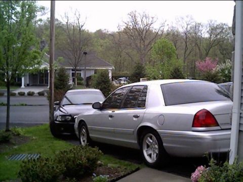 2002 Ford Crown Victoria - A silver 2002 Ford Crown Victoria is parked next to a dark sedan on a street with houses and trees in the background.