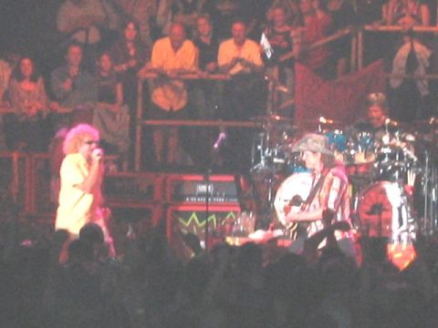 1989 Ford Bronco - Wide concert shot of a curly-haired singer in a yellow shirt at the microphone on the left, a guitarist in a bucket hat by a white drum kit…