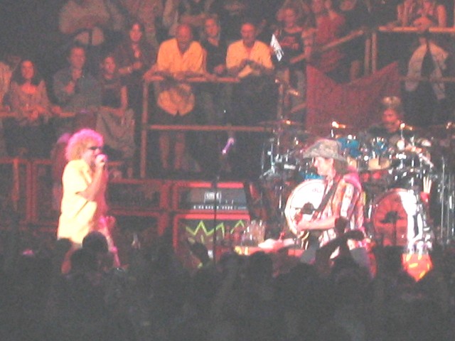 Concert stage with singer in yellow shirt at mic, guitarist, drum kit and amp stacks