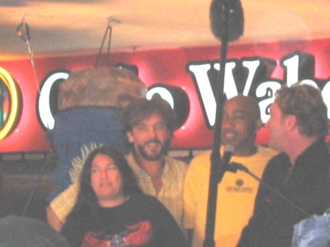 1989 Ford Bronco - Four people stand together on a stage in front of a large illuminated red sign and a microphone on a boom stand, one wearing a yellow shirt…