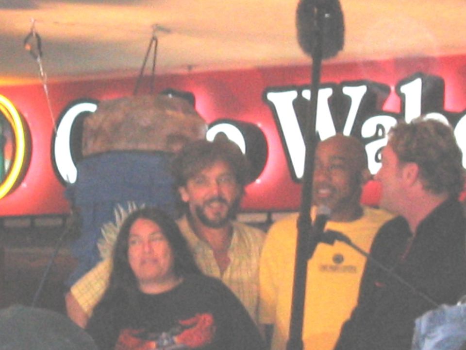 Four people on stage in front of a lit red sign and microphone at a club event