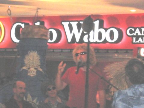 1989 Ford Bronco - A curly-haired man in sunglasses and a red T-shirt sings into a microphone beneath an illuminated red "Cabo Wabo" sign, with a decorated ba…