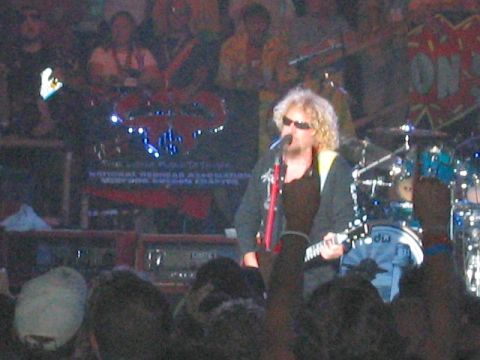 1989 Ford Bronco - A blond curly-haired singer in sunglasses and a gray jacket plays a red guitar at the microphone on a concert stage, with a drum kit, ampli…