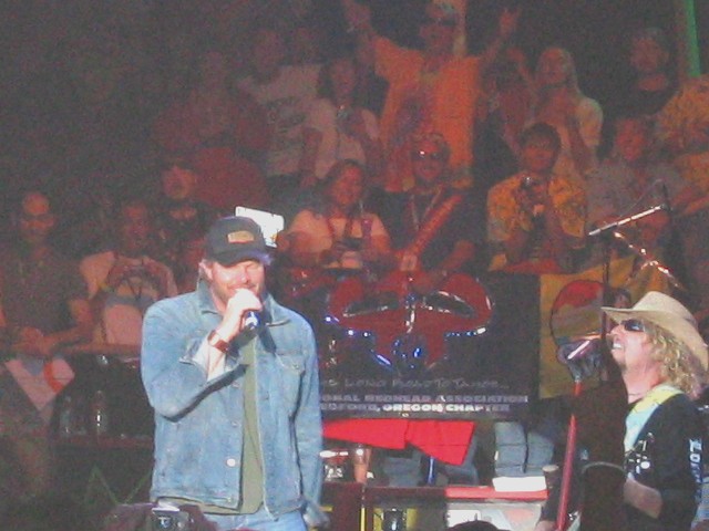 Bearded singer in denim jacket at microphone on stage with guitarist in straw hat