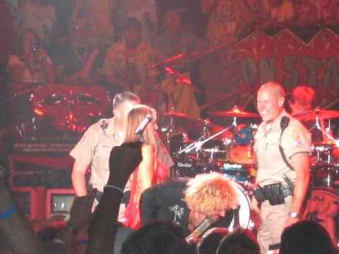 1989 Ford Bronco - Concert stage scene with two uniformed officers in tan shirts standing beside a woman in a red top holding a microphone, with a red drum ki…