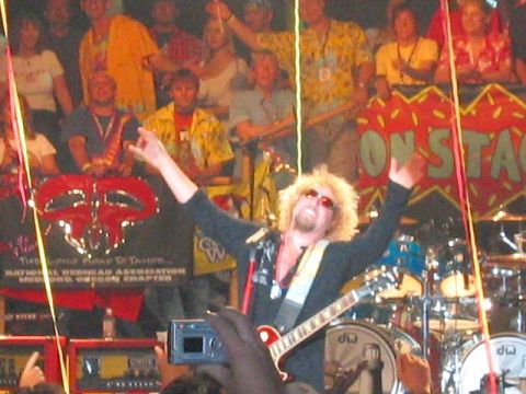 1989 Ford Bronco - A blond, sunglasses-wearing rock guitarist in a dark jacket raises both arms on stage in front of a drum kit and a crowd of onlookers, with…