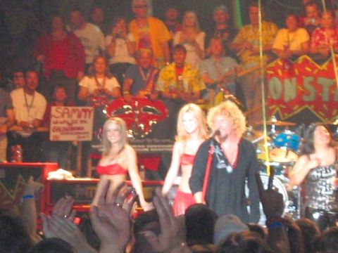 1989 Ford Bronco - A live concert stage with a blond singer in a black shirt at the microphone, backup dancers in red outfits, a drum kit, and a red banner re…
