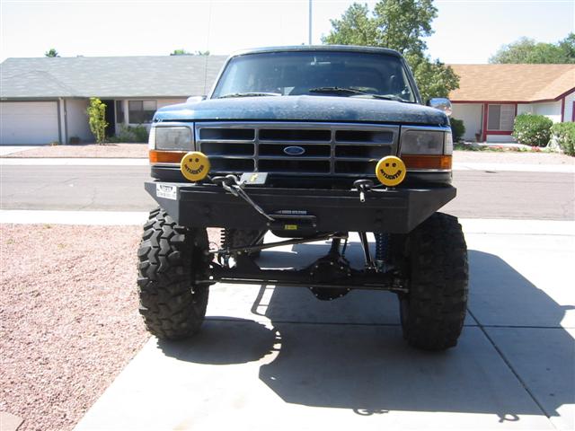 Lifted dark blue Ford Bronco, front view, big tires and smiley-face lights on bumper.