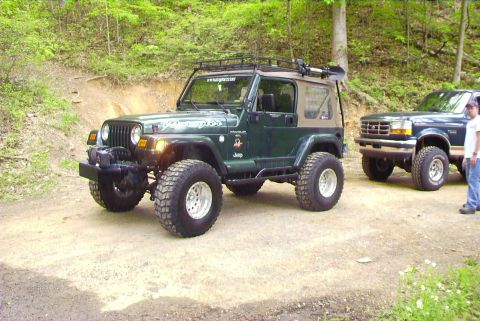 1995 Ford Bronco - Two off-road vehicles are parked on a dirt path surrounded by trees.