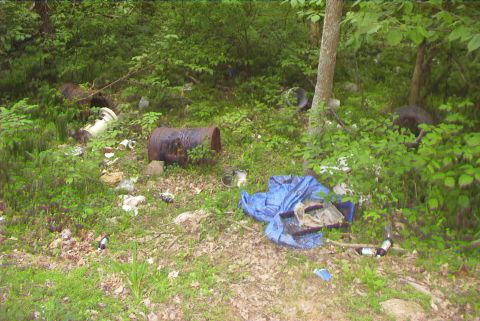 1995 Ford Bronco - A wooded area is littered with trash including barrels, a blue tarp, and discarded bottles.