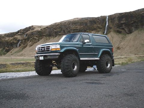 1996 Ford Bronco - Modified 1996 Ford Bronco with large off-road tires sits near a scenic waterfall landscape.