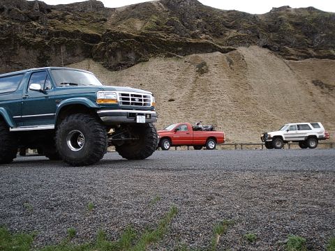 1996 Ford Bronco - A lifted 1996 Ford Bronco with massive tires is parked alongside a red pickup truck and a white SUV on a gravel road with a mountainous bac…