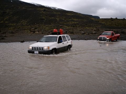 1996 Ford Bronco - Two SUVs navigate a river crossing in a , mountainous landscape under an overcast sky.