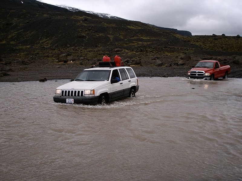 White Jeep SUV and red Dodge pickup crossing a muddy river in a volcanic landscape