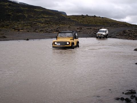 1996 Ford Bronco - Two off-road vehicles navigate a shallow river in a , mountainous terrain under an overcast sky.