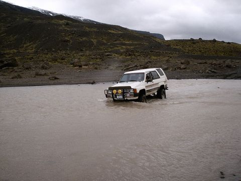 1996 Ford Bronco - A white SUV navigates a shallow river in a rocky, barren landscape under an overcast sky.