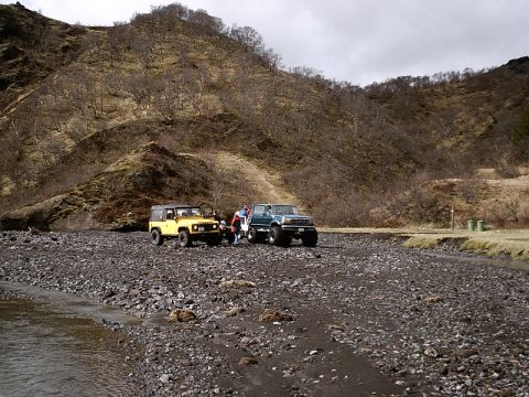1996 Ford Bronco - Two off-road vehicles are stopped on a gravelly riverbed with a barren, hilly landscape behind them.