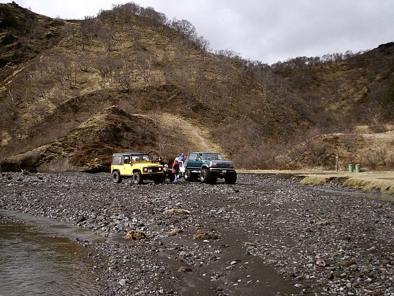 Green lifted Bronco and yellow Land Rover parked on rocky riverbed near hills