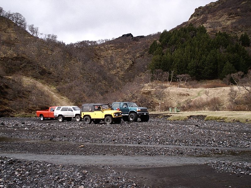 Four 4x4 vehicles including a yellow soft-top and teal SUV parked on a rocky riverbed in a valley