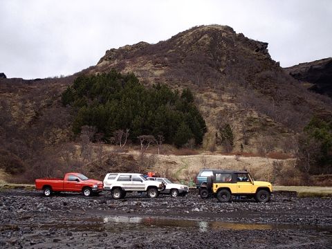 1996 Ford Bronco - A convoy of off-road vehicles, including a red pickup and a white SUV, navigate a rocky, wet landscape under a cloudy sky.
