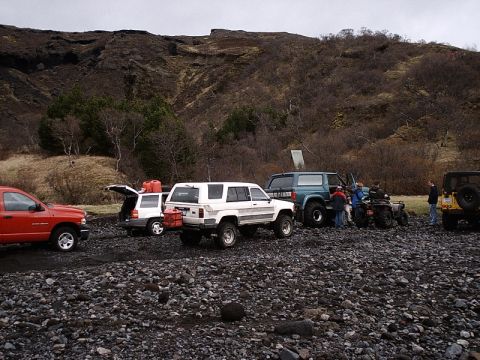 1996 Ford Bronco - A group of off-road vehicles are gathered on a rocky beach, with people milling around and gear loaded in some of the vehicles.
