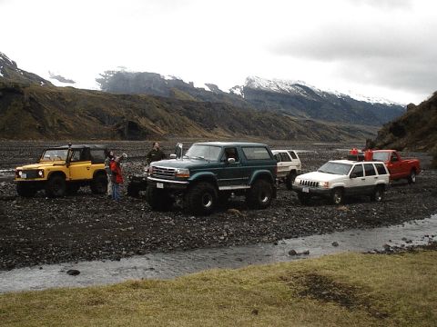1996 Ford Bronco - Several off-road vehicles, including a 1996 Ford Bronco, are parked in a rocky riverbed with snow-capped mountains.