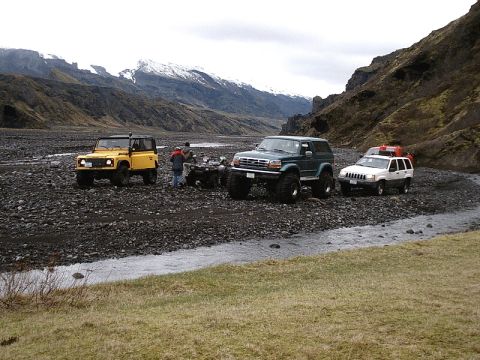 1996 Ford Bronco - Several off-road vehicles are parked in a rocky riverbed, with a stream flowing nearby and mountains in the distance.