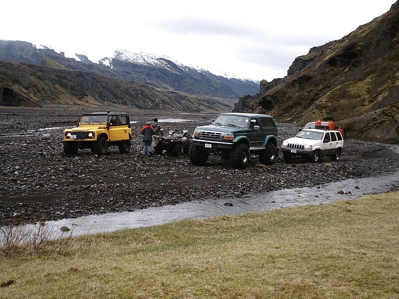 Green lifted Ford Bronco among off-road vehicles in a rocky river valley with snowy mountains