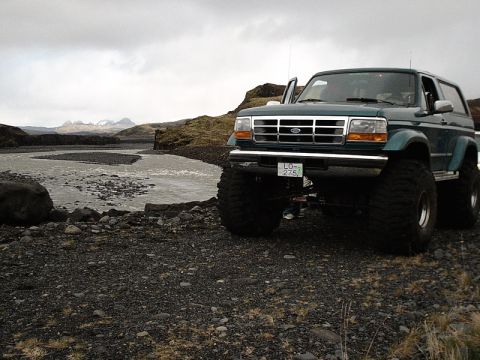 1996 Ford Bronco - This modified 1996 Ford Bronco, massive tires, is parked in a , remote landscape with a river and mountains in the background.