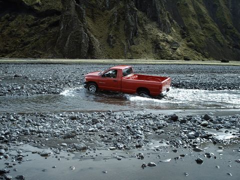1996 Ford Bronco - A red pickup truck navigates a rocky riverbed, kicking up water as it drives through the shallow stream.