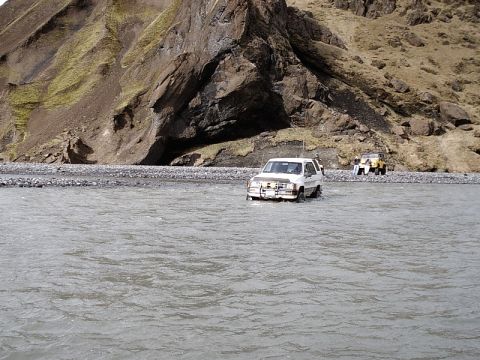 1996 Ford Bronco - A white SUV navigates a river, followed by a yellow vehicle, with a rocky mountainside in the background.